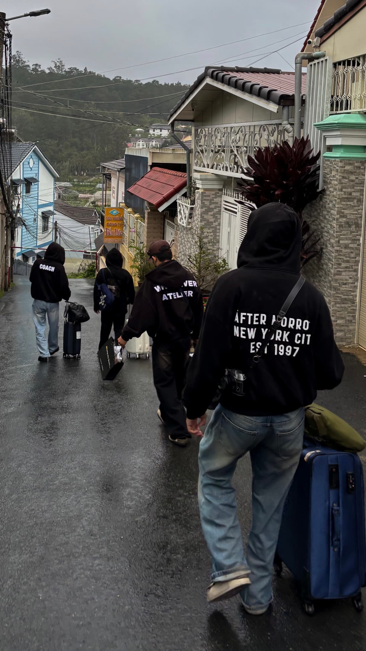 Group wearing black hoodies wheeling luggage down a wet hillside street